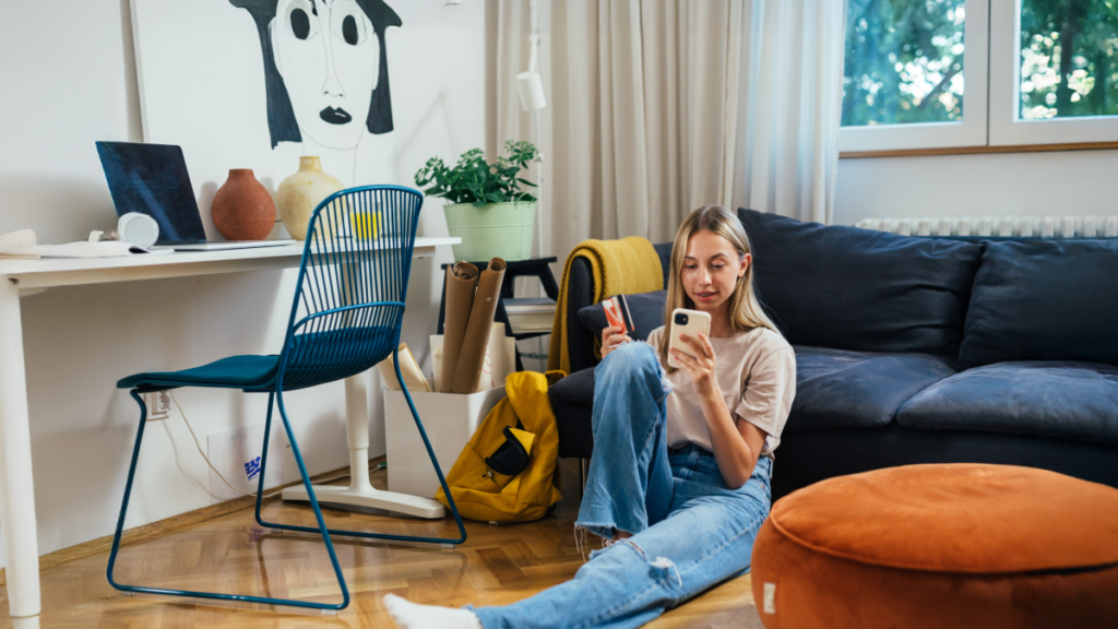 Young woman sitting comfortably on the floor at home, leaning against a couch with a mobile device in one hand and a credit card in another.